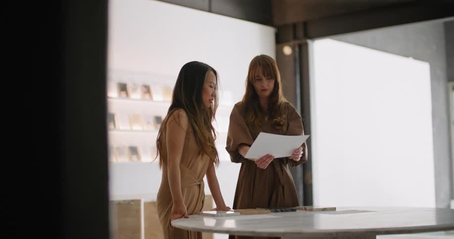 Two women standing over a table looking at a paper and pointing and talking
