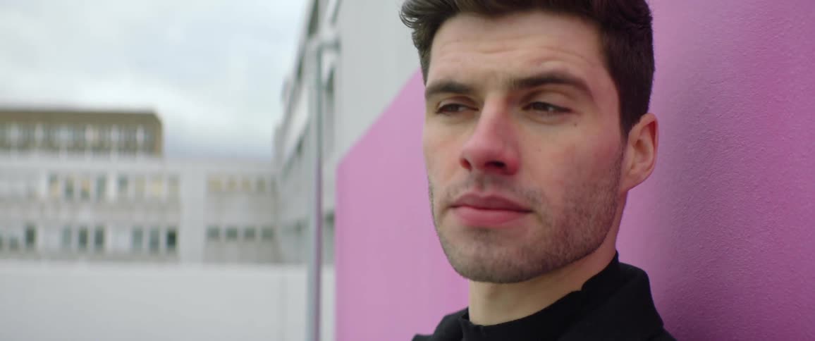 A close up of a mans face looking contemplatively while standing against a pink wall outside