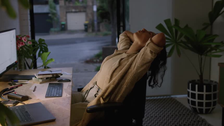 A woman is sitting at a work desk at dusk while yawning and stretching