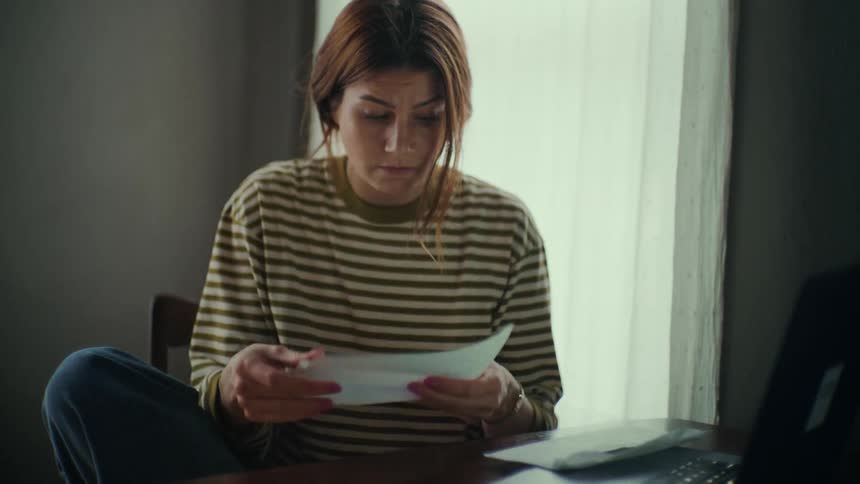 A woman unfolding a paper as she sits at a desk in front of a laptop