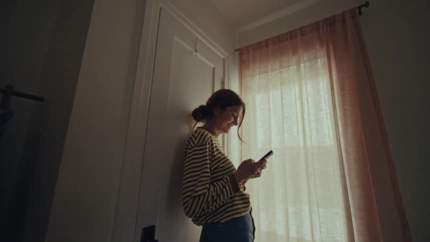 A woman looking at her phone laughing standing in front of a window