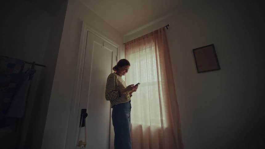 Woman with striped shirt and jeans uses mobile phone in room next to window