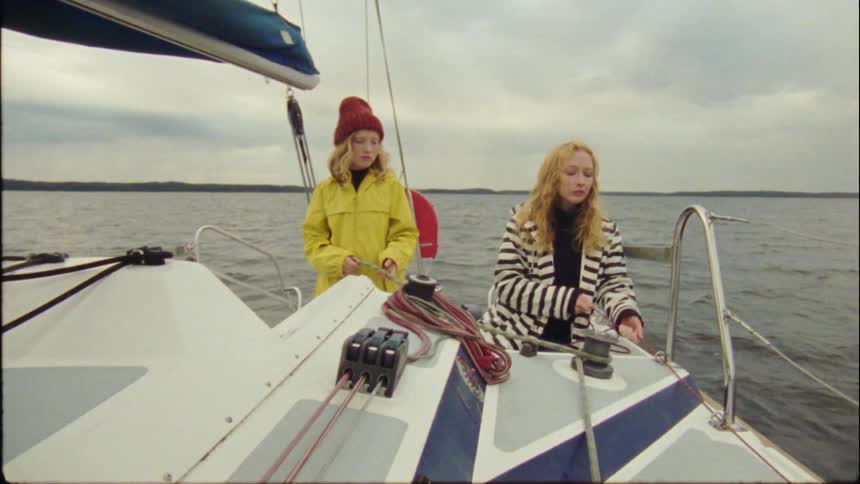 Blonde mother with wavy hair and daughter in red hat and yellow coat pull rope on boat on a cloudy day