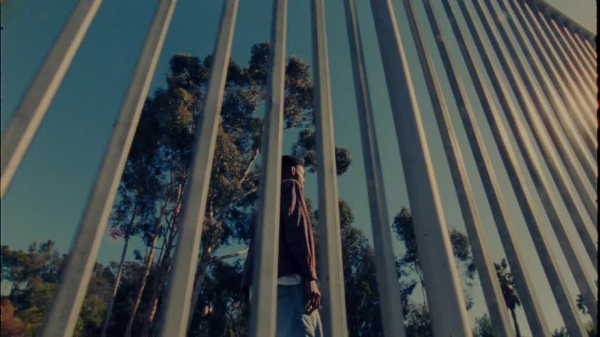 Man stands holding a skateboard near metal bar fence on sunny day