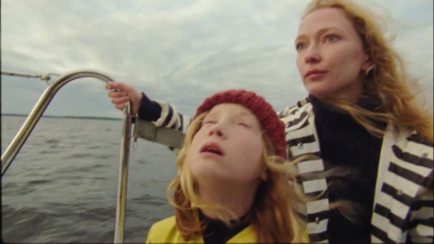 Blonde mother with wavy hair and striped jacket sits next to daughter with red hair and yellow coat traveling on boat on cloudy day