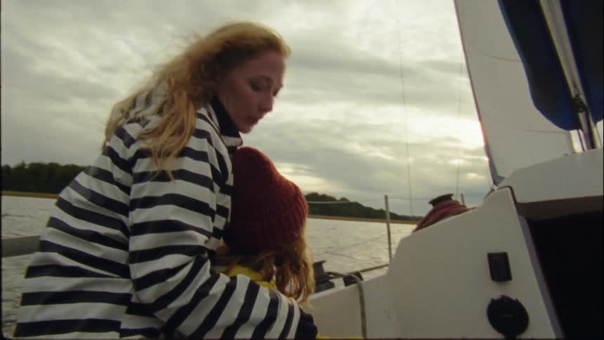 A blonde woman puts her arm around her young daughter while sailing under cloudy skies