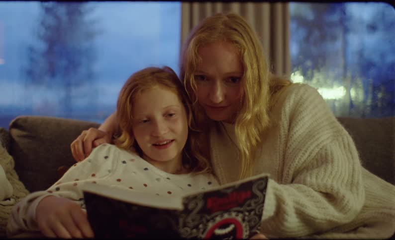 Mother and daughters sit on couch reading a book together as rain falls outside