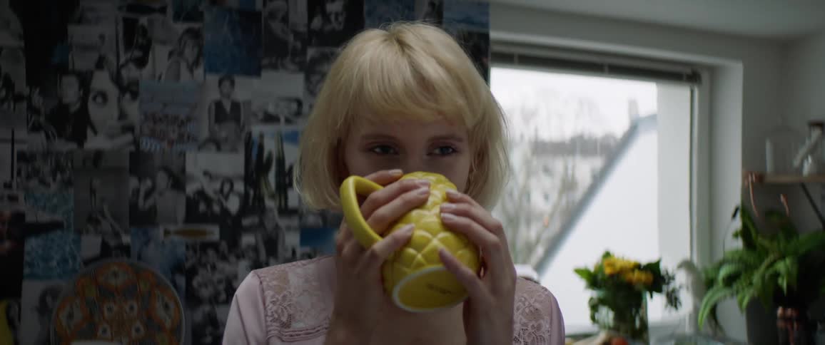 A young blonde woman is drinking from a yellow mug while at home