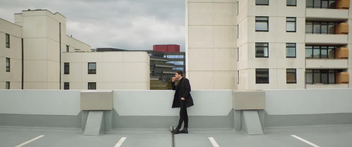 A man standing against a half wall in a parking lot with his hand on his chin
