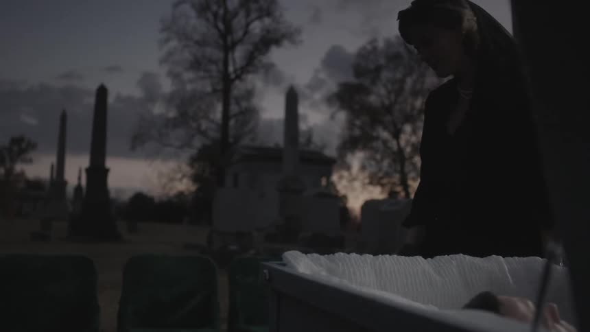 A woman walks away from a casket at dawn