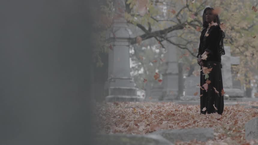 Leaves fall on a woman standing in a cemetery