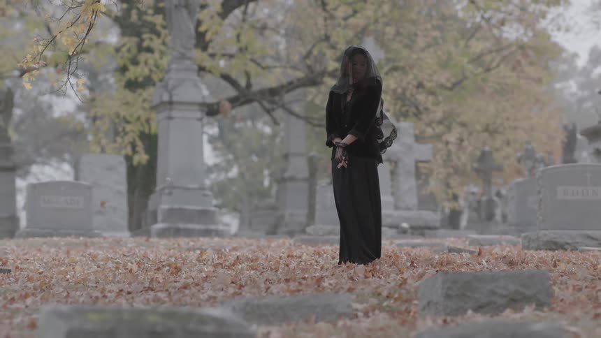 A veiled woman stands in a cemetery as leaves fall around her