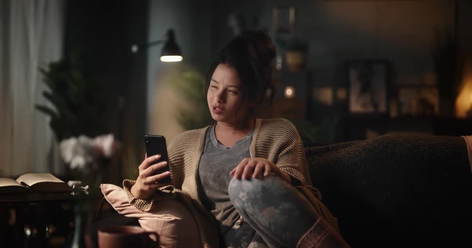 A woman sits on her couch at home and holds her cell phone as she talks to someone on it