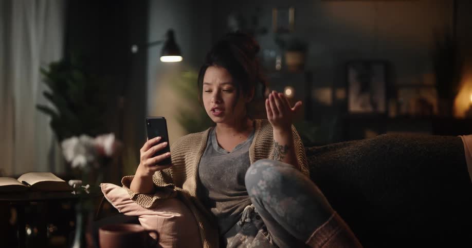 A woman is sitting on the couch holding her phone out as she smiles and laughs as she talks and moves her hands
