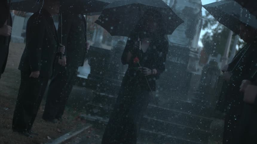Mourners in black clothing carrying umbrellas stand in a cemetery in the rain