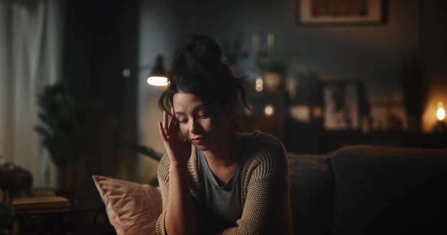 A woman is sitting on the couch in front of a laptop computer as she speaks in a video conference and closes the computer resting her chin on her hand and looking sad
