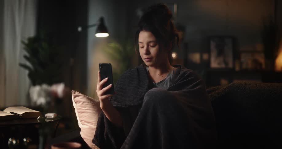 Young woman sitting on couch covers herself with blanket and facetimes with someone on smartphone