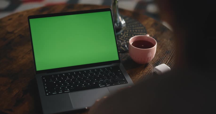 A woman at home sits in front of a laptop with a green screen
