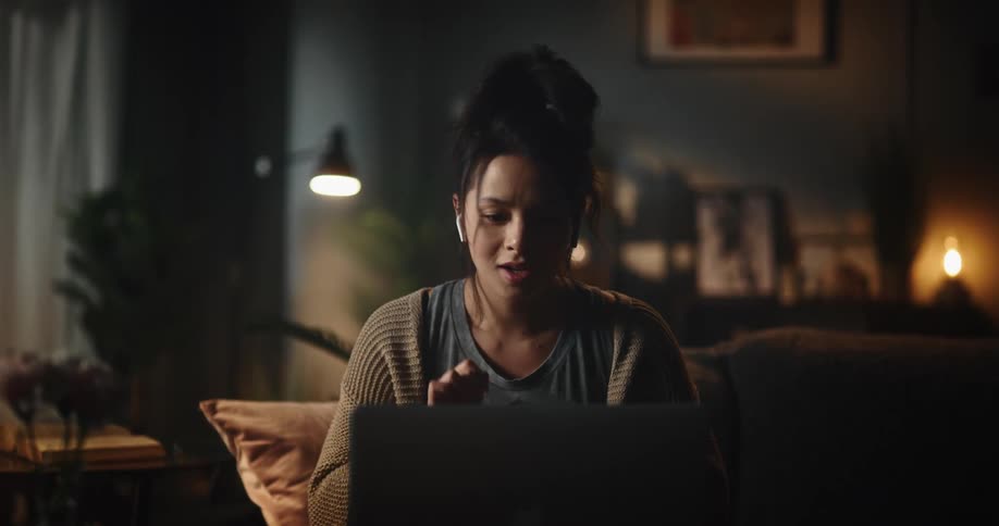 A woman walking in her living room sitting on her couch with her laptop and putting ear buds in her ear