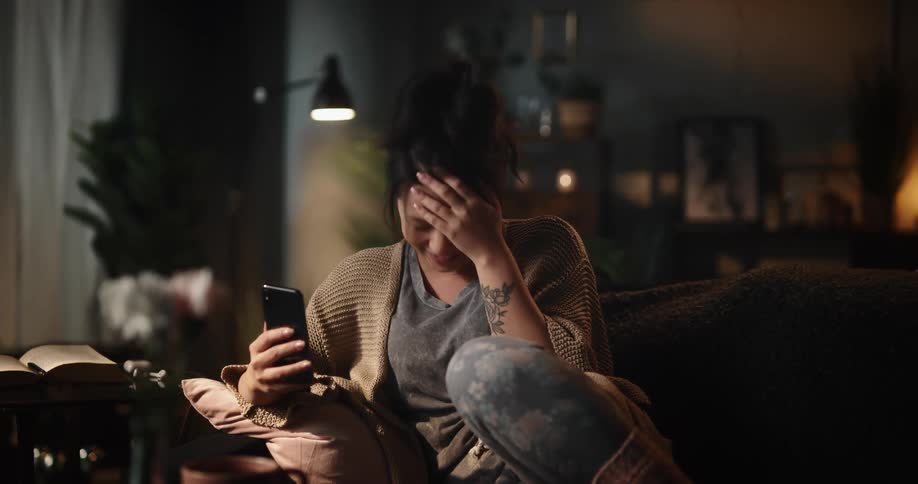Young brunette woman lounging on couch and facetiming with someone on smartphone laughing and talking