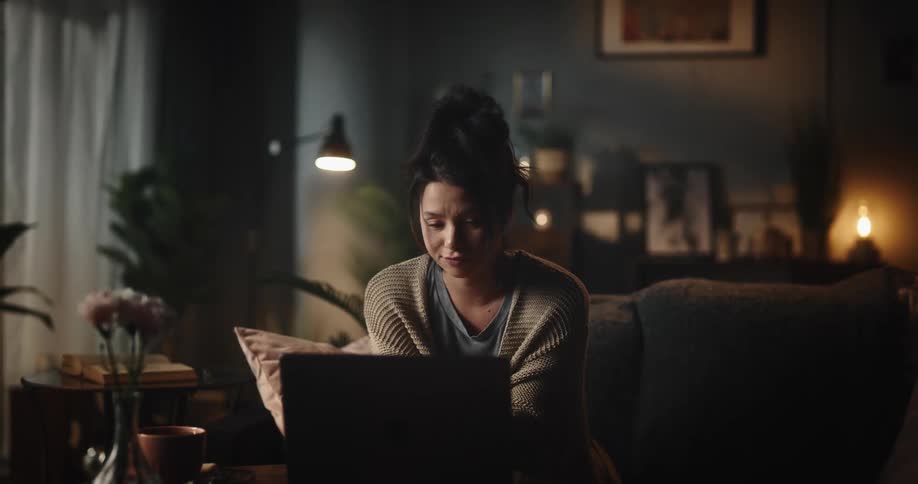 A woman is sitting on the couch in front of a laptop computer as she moves her hands and shakes her head as she speaks in a video conference
