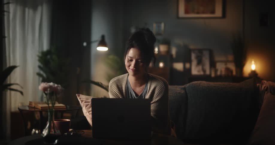 A woman is sitting on the couch as she opens her laptop computer and puts earbuds in her ears as she smiles and has an online conversation