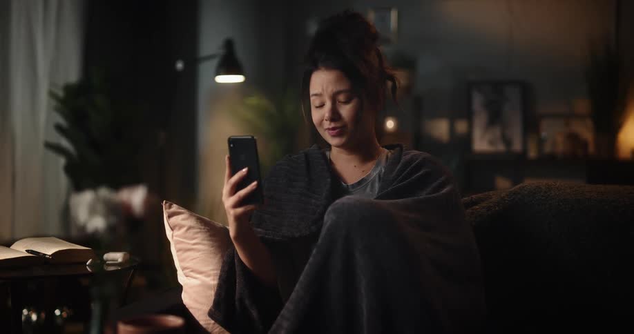 Woman sitting on couch tearfully frowns while talking via video call on a smartphone