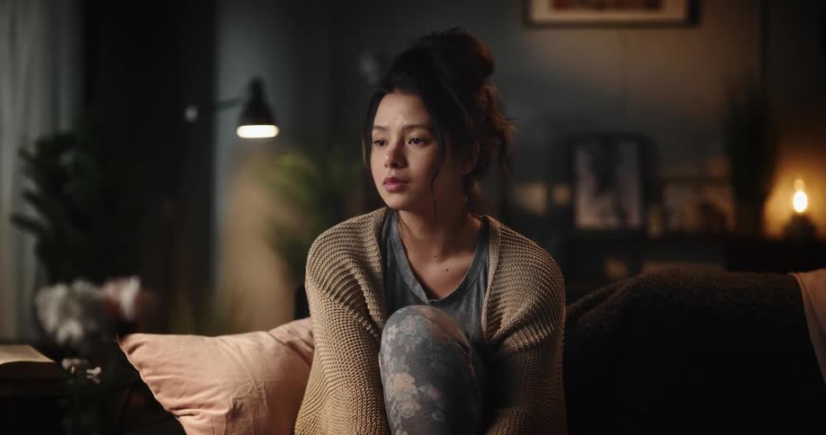 Young woman sitting on couch deep in thought worry and close to crying looks at cam