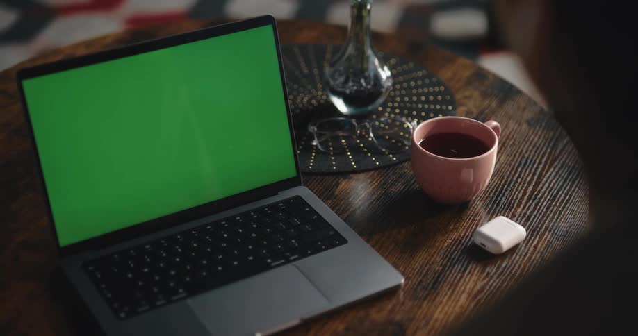 A laptop with green screen a pink cup with coffee in it and a person picking up ear buds off the table