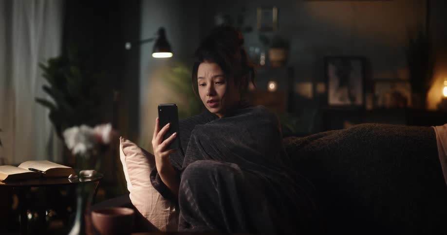 A woman is sitting on the couch holding her phone out as she talks and appears to be sad