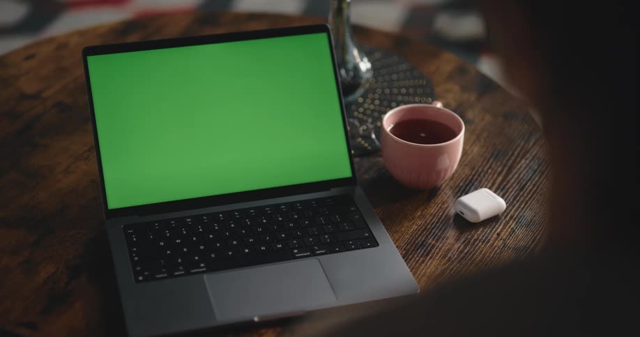 A woman sits at home in front of a laptop with a green screen and moves her hands around and puts earbuds in her ears