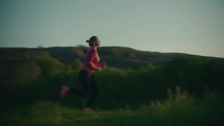 A woman is jogging through a field near mountains as the sun shines brightly on the horizon