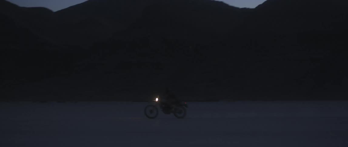 A dirt bike with a single head light on driving across the desert at night with mountains in the background