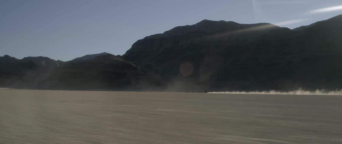 A motorcycle speeds across the desert along a mountainside and leaves a trail of smoke behind