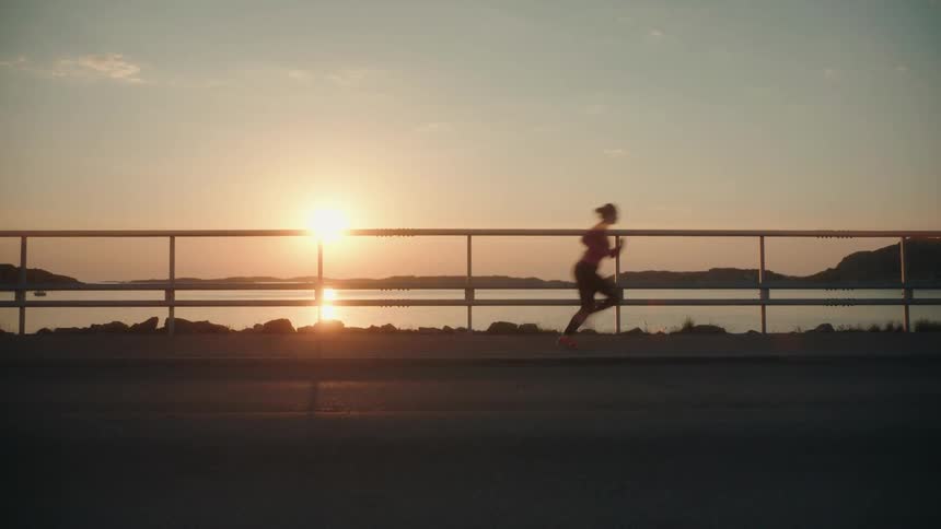 A young woman is jogging along a sidewalk that overlooks a large body of water at sunset