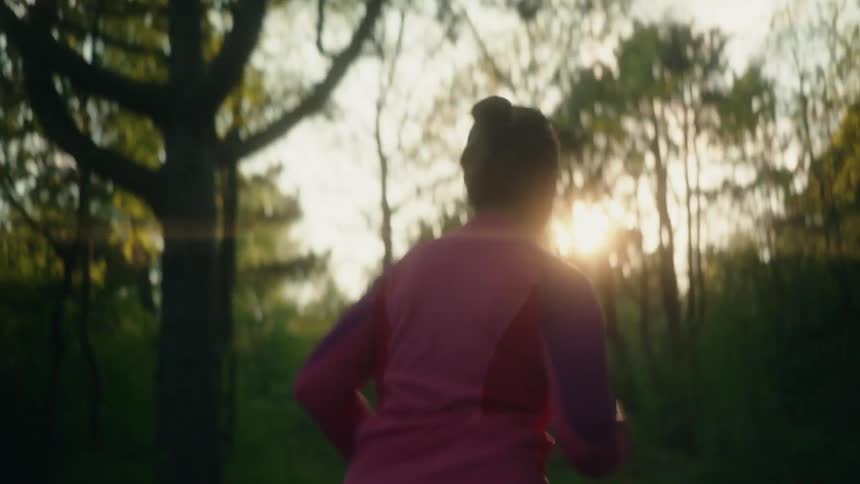 A person wearing a pink top runs through a forest with the sun ahead of them