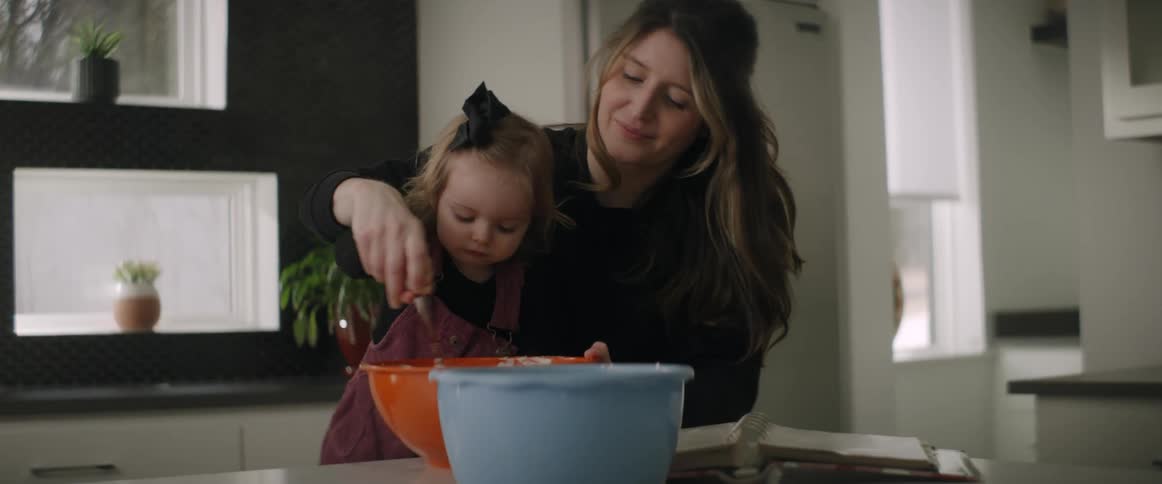 A mother helps her daughter mix ingredients with a spoon in an orange bowl