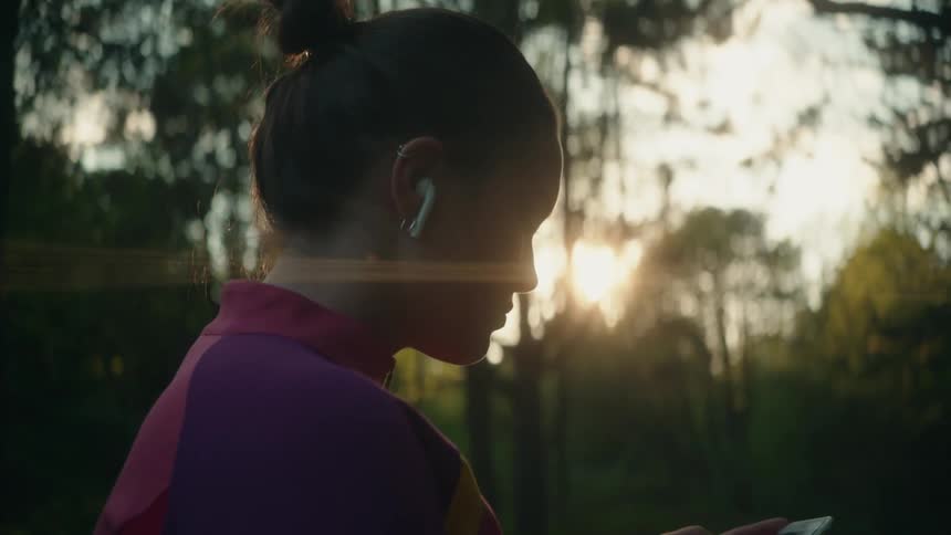 A woman is standing in the woods as she turns her ear bud and looks at her phone then she walks into the field