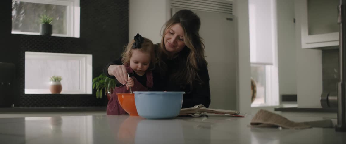A woman has her toddler daughter help her stir food in a bowl