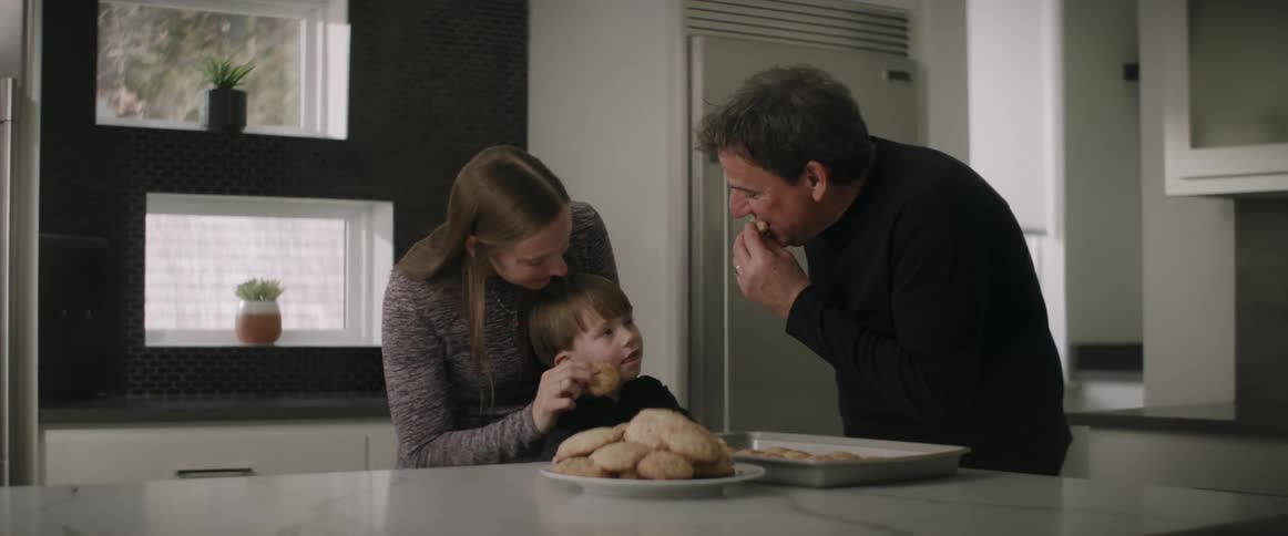 Man in black sweater and woman with fair hair and boy eating cookies in the kitchen
