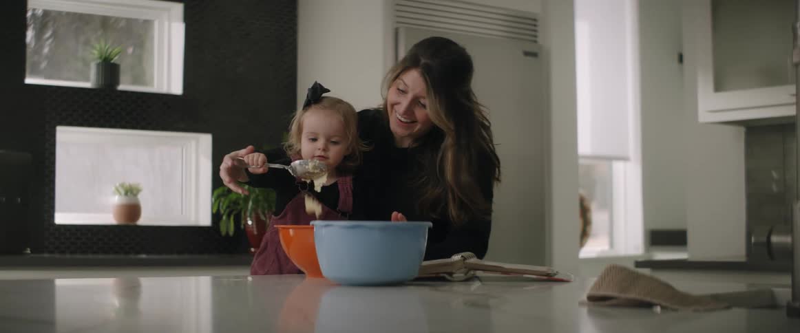 A mother helps her daughter mix with a spoon as she laughs