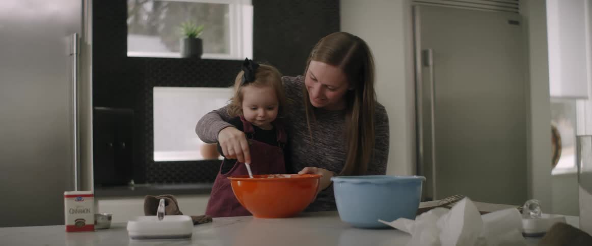 Mother with brown hair and sweater stirs in red bowl holding daughter in the kitchen