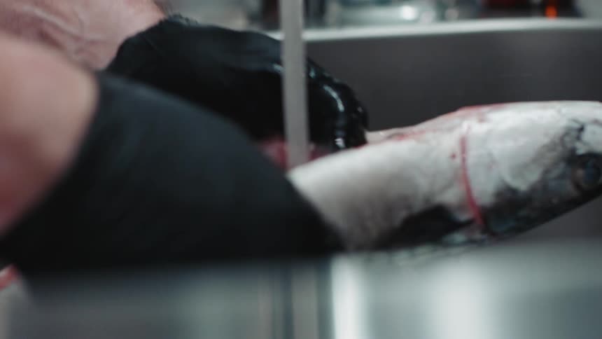 Chef wearing black latex gloves cleans a gutted fish under a stream of water in the kitchen sink