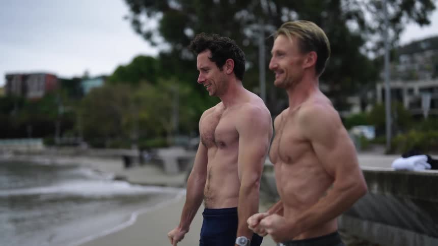 Two men are standing bare chested on a beach and they appear to be cold as they walk toward the water