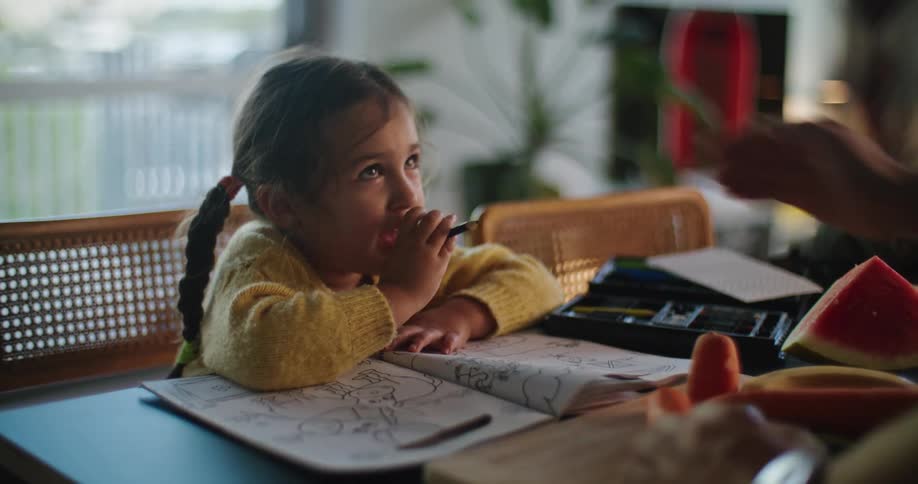 A young girl sits at table with a coloring book and her mom reaches across and wipes her hair off her forehead