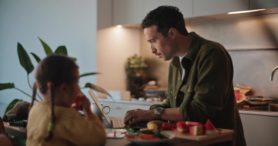 A man typing on his laptop as he talks to his daughter sitting at the counter eating