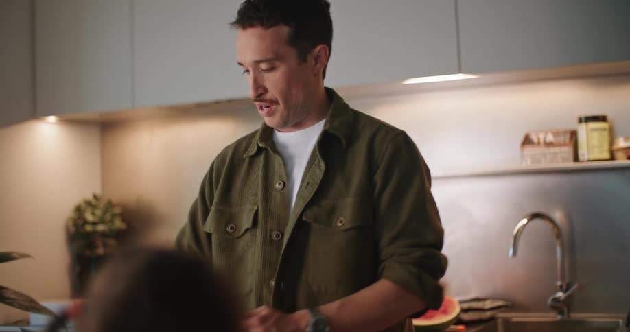 A man turning around in his kitchen with a cup in his hand talking to someone sitting in front of him