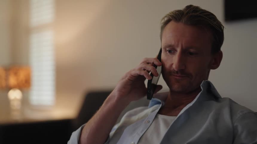 A man sitting on the sofa while speaking on the smartphone turns off the television and then picks up a piece of paper with his left hand