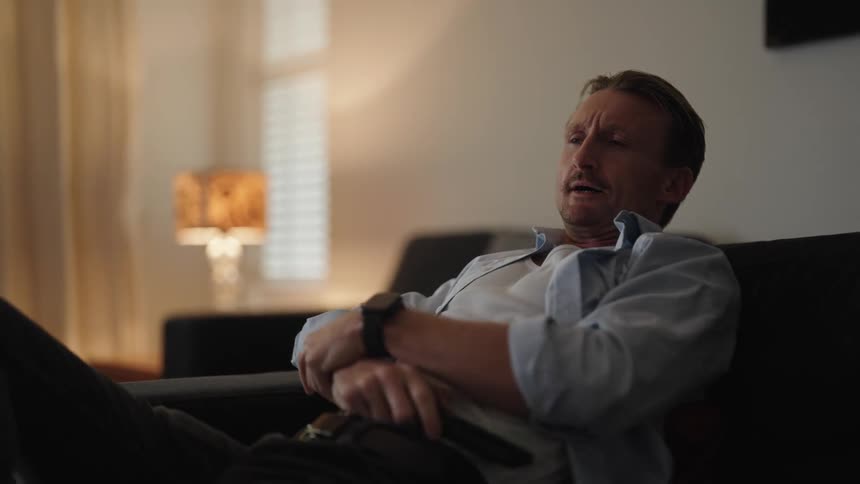 Tired man yawns and reclines on a couch at home while watching television