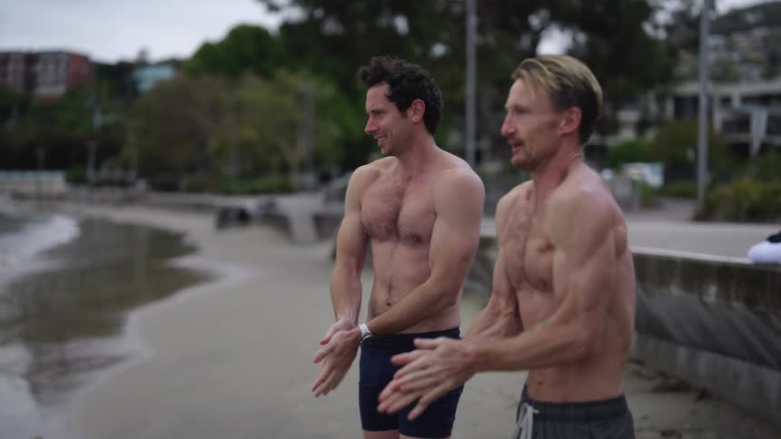Men wearing swim trunks greet each other on beach and try to stay warm